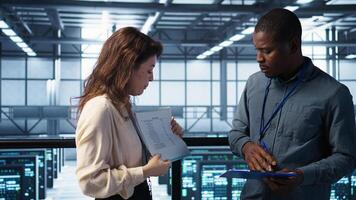 IT professionals looking over data center checkup checklist paperwork. African american man and colleague collaborating in server farm reading documentation files, camera B photo