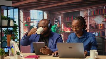 Black woman working while partner listens to music next to her, enjoying time at home together. Freelancer solving daily tasks on laptop, boyfriend scrolling web. Camera A. photo