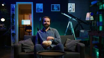 Man turning on TV using remote control, enjoying leisure time while munching on popcorn. Couch potato person in neon illuminated apartment eating salty snack, entertained by television, camera B photo