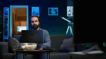 Man watching TV in apartment office and solving business tasks on laptop, working from home. Freelancer on couch in living room looking at movies and working on device, camera B photo