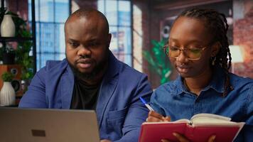 Black employees organizing calendar appointments using laptop and ledger, checking papers and synchronizing important tasks on schedule. Man and woman review files at home. Camera A. photo