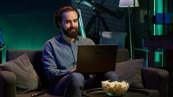 Man typing on laptop in neon lit apartment, sitting on couch and watching TV with bowl of popcorn ready. Teleworking person solving tasks on notebook with film playing in background, camera A photo