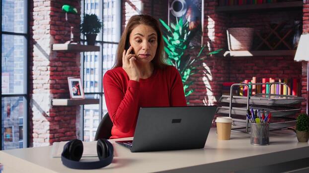 Woman in home office talking on mobile phone while working on laptop. Remote worker juggles calls, meetings and inquiries, using communication skills and creativity to solve tasks. Camera B. photo
