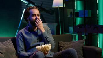 Amused person sitting on couch using TV, binge watching comedy series on streaming services and enjoying popcorn. Man laughing at funny shows on television, eating snack, camera A photo