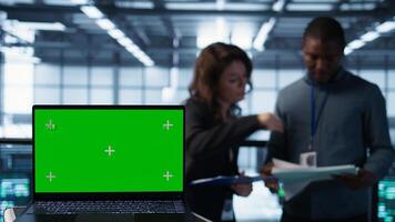 Green screen laptop used by data center admins in background reviewing equipment diagnostics. Chroma key notebook used by coworkers adjusting server room configurations, camera B photo