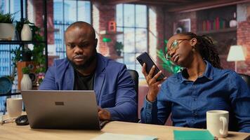 African american people spending time together at home with different activities, young man working and earning money while girlfriend uses smartphone, scrolling on websites. Camera A. photo