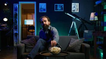 Man taking seat on sofa, watching television and getting entertained in relaxed home. Person arriving in apartment sitting down on couch to enjoy movies, turning on TV, eats popcorn, camera B photo
