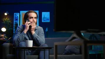 Man appalled by upsetting breaking news story on TV, on phone call with family member. Person at home scared by troubling events broadcasted on TV, calling friend using smartphone, camera B photo