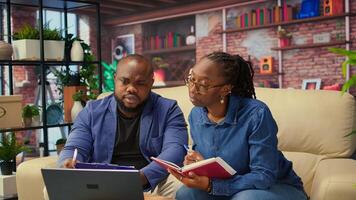 Black people overseeing schedule and planning activities at coffee table, working on time management with online tools. Couple utilizing notebooks and taking handwritten notes. Camera A. photo
