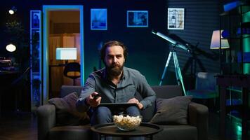 Man using laptop and watching breaking news on TV in neon lit apartment. Person turning volume on television, surprised by events while sitting on couch using notebook, camera B photo