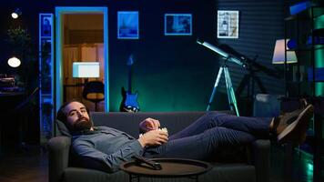 Smiling man on couch eating popcorn and watching movie on streaming service, enjoying time off. Upbeat person laying down in apartment consuming snacks and looking at films, camera B photo