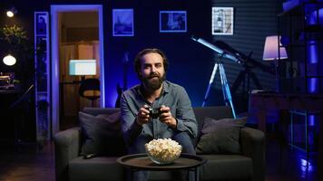 Excited man holds controller in neon illuminated apartment, playing videogames. Happy gamer on couch using joystick, participating in PvP game on console system, relaxing and having fun, camera B photo