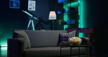 Bowl of popcorn on coffee table in front of cozy couch in empty neon lit apartment. Close up shot of snack prepared for movie night in home theatre used for entertainment sessions photo