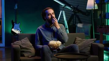 Man turning on TV using remote control, enjoying leisure time at home, munching on popcorn. Couch potato person in apartment eating salty snack, entertained by television, camera A photo