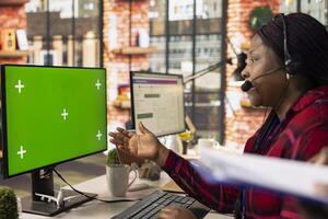 Call center agent talking in headset mic, using green screen mockup PC, providing step by step guidance to customer. Customer center employee at chroma key computer walking clients through solutions photo