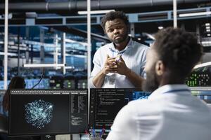 Server farm manager supervising engineer looking at data analysis graph to reduce failures. Data center team leader overseeing worker looking at AI visualization dashboard to pinpoint gear glitches photo