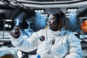 Female astronaut doing videocall, talking with family, presenting space station. African american woman in spaceship using smartphone to recording herself sharing mission progress with loved ones photo