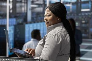 Smiling technician in server room using laptop to implement and manage data backup solutions. Happy data center engineer identifying and resolving technical issues, ensuring efficient recovery photo