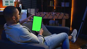 Black remote worker looking at green screen on his device at home, relaxing in the living room and solving freelancing tasks. Guy being casual in trendy apartment, teleworking. Camera A. photo