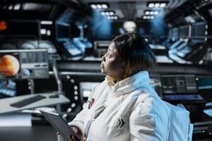 Woman in orbital station analyzing Mars planetary information on tablet. African american spaceship crew member doing Martian surface research using device to assist future planetary exploration photo