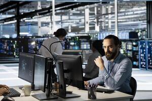 Technician using PC in data center to update server tech, configuring equipment using software tools. Server room employee reviewing hardware analytics, ensuring reliable network operations photo