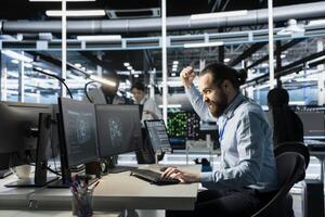 Angry software developer in data center clenching fists while looking at AI visualization interface on monitor. Frustrated technician reacting to artificial intelligence software glitches photo