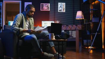 Black freelancer sitting on sofa and reading some papers to do research, multitasking with archives in the living room. Adult diving into professional or personal topics of improvement. Camera A. photo