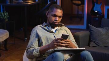Black man taking a break from work by scrolling his smartphone at home, messaging friends on social media and having fun. Guy relaxing in stylish living room with neon lights. Camera B. photo