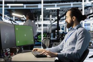 Engineer in data center using green screen mockup PC to update storage arrays software, configuring network virtualization. Man in server room using chroma key computer to verify settings photo