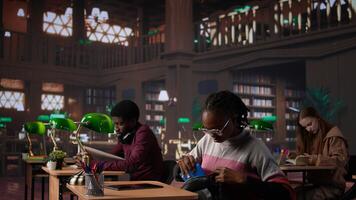 Black girl focuses on exam preparations in campus library, takes notes on laptop from reference books in peaceful study setting. Girl uses general database materials. Camera A. photo
