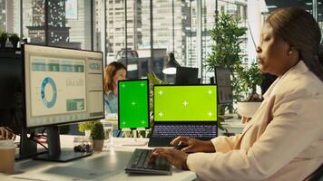 Green screen laptop and tablet on desk next to employee editing documents on computer. Chroma key devices next to worker analyzing data charts, interpreting statistical information, camera B photo