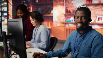 Smiling call center agent speaking with unhappy customers, using audio headset at office desk. Customer support worker handling clients complaints in company workspace, camera B photo