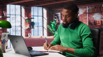 African american teleworker writing personal notes at desk in modern loft, remote job. Productive guy sitting at home office, using pen to take notes in textbook. Camera B. photo