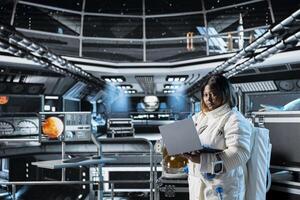 Female cosmonaut monitoring system diagnostics on laptop before Mars expedition. African american astronaut in spaceship explores red planet to support human life beyond Earth photo