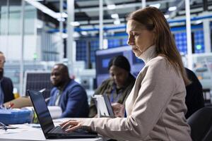 Factory employee works on laptop next to executives discussing solar panel automation strategies. Worker typing on notebook next to management team enhancing sustainable manufacturing processes photo