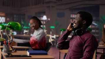 Noisy black guy disturbing pupils at library by speaking very loudly, being rude and answering phone call in study area. Disruptive annoying man asked to keep silence. Camera A. photo