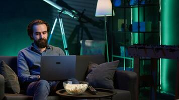 Man watching TV and solving business related tasks on laptop, working from home. Freelancer on couch in living room looking at movies and working on notebook device, camera A photo