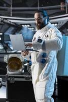 Astronaut researcher verifying mission parameters using laptop, carrying out research aboard. Scientist wearing cosmonaut gear in spaceship updating flight log using notebook device photo