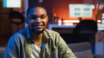 Portrait of black freelancer laying on the sofa and texting on phone, relaxing and smiling in his modern loft. Man solving daily teleworking tasks from the living room with cool lights. Camera B. photo