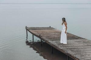 unrecognizable woman in white outfit stands on wooden pier, calm lake, peaceful overcast, viewed from behind with contemplative mood, mindfulness, travel retreats, wellness photo