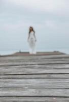 blur woman silhouette on wooden pier, calm sea, dressed in white outfit under a pale cloudy sky, mindfulness, wellness retreat, travel blog photo