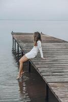 unrecognizable woman in a white dress, sitting on the edge of wooden pier, dipping her feet into calm water with a light splash, gazing at horizon under soft cloudy sky, relaxation, travel, vertical photo