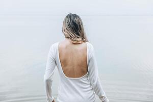 unrecognizable woman standing with back exposed in open white dress near calm lake, facing the water with relaxed posture and serene mood, concept of wellness, spa, fashion editorial photo