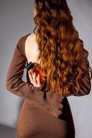 woman in brown dress with long red wavy hair holding perfume bottle behind her back, fashion studio shoot with soft shadows, back view, fragrance branding, beauty marketing, cosmetics retail photo