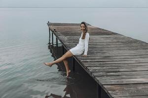 Asian woman in a white dress, sitting on the edge of wooden pier, dipping her feet into calm water with a light splash, gazing at horizon under soft cloudy sky, relaxation, travel photo
