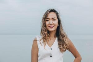 Asian woman smiling and looking at camera on a calm lakeside, dressed in white outfit, displaying a confident and cheerful emotion, concept of wellness, skincare, personal development photo