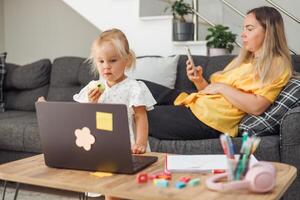 Young blond European girl eating apple with laptop on table. Mother woman relaxing on couch using smartphone. Home environment. photo