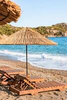 Wooden loungers with straw umbrellas line the sandy beach while gentle waves lap at the shore under a clear blue sky. photo