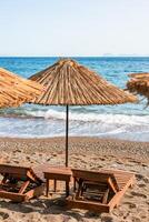 Wooden lounger with straw umbrellas line the sandy beach while gentle waves lap at the shore under a clear blue sky photo