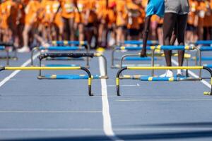 Several hurdles placed on a blue running track with a group of athletes in orange shirts waiting behind photo